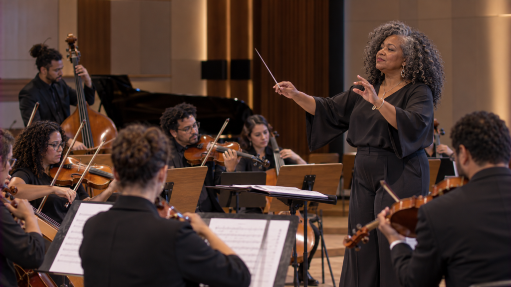 Maestrina brasileira negra de meia-idade conduzindo uma orquestra durante ensaio.